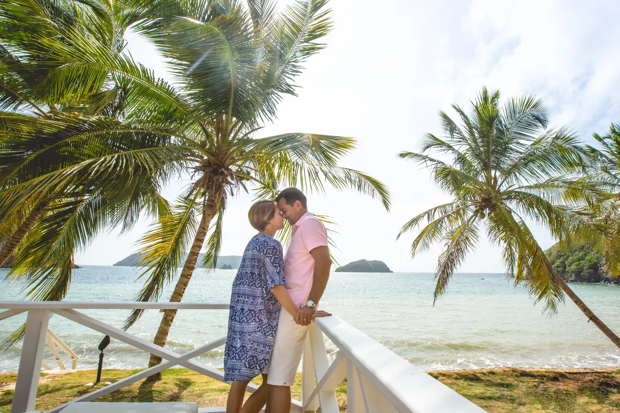 A couple sharing a kiss by the railing under the palms, with the sea behind