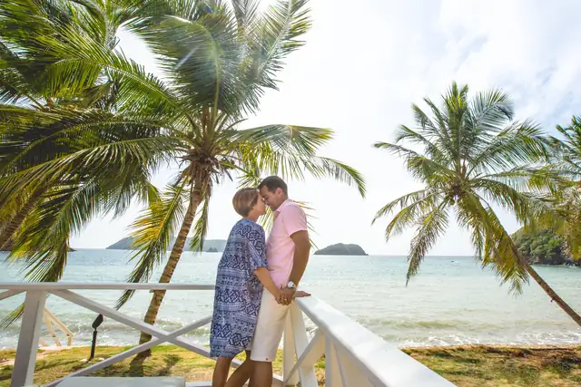 A couple sharing a kiss by the railing under the palms, with the sea behind