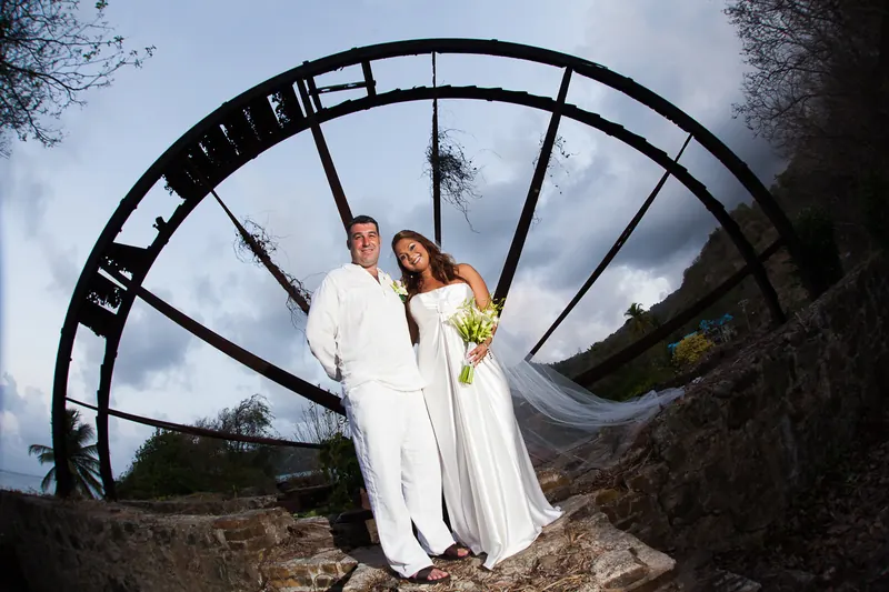 A bride and groom in white beneath the arch of an old water wheel