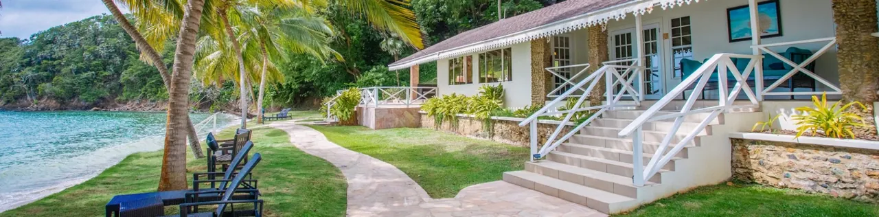 A beachfront bungalow with white railings on the lawn beside the bay, with palms and loungers along the shore